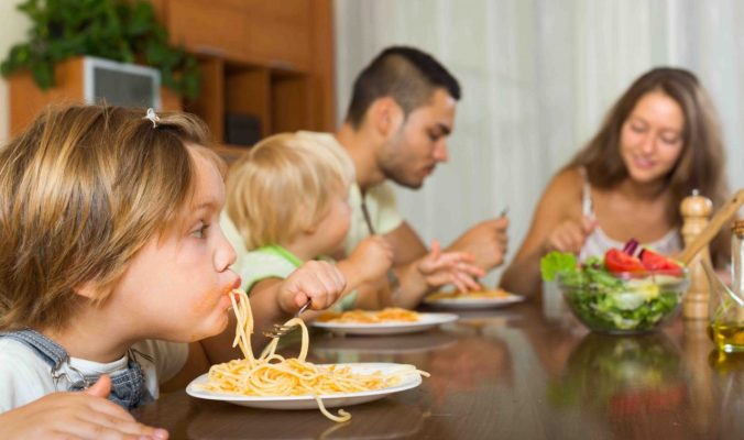 Happy family with playful little daughters eating with spaghetti at table. Focus on girl