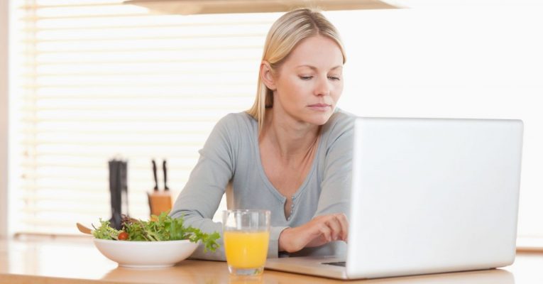 Young woman with salad and orange working on laptop in the kitchen
