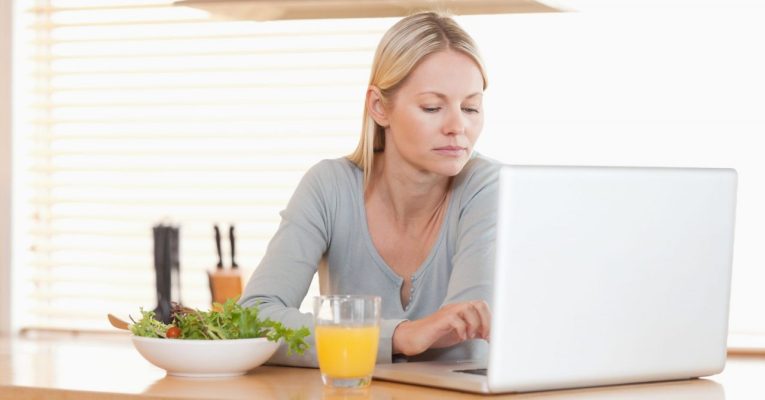 Young woman with salad and orange working on laptop in the kitchen
